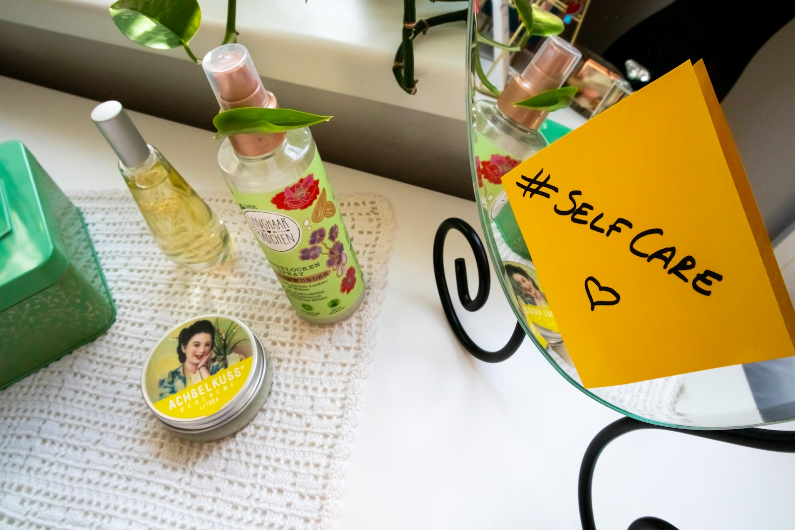 A neatly arranged setup on a white table includes two bottles of spray, one with floral designs, a tin with vintage-style artwork, and a green box. A yellow sticky note with '#SelfCare' and a heart is attached to a mirror. Green leaves add a touch of nature to the scene.